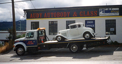 Photo of past Totem Towing deck truck branded with Audy Towing, towing a classic car on its deck in front of Audy Autobody & Glass.